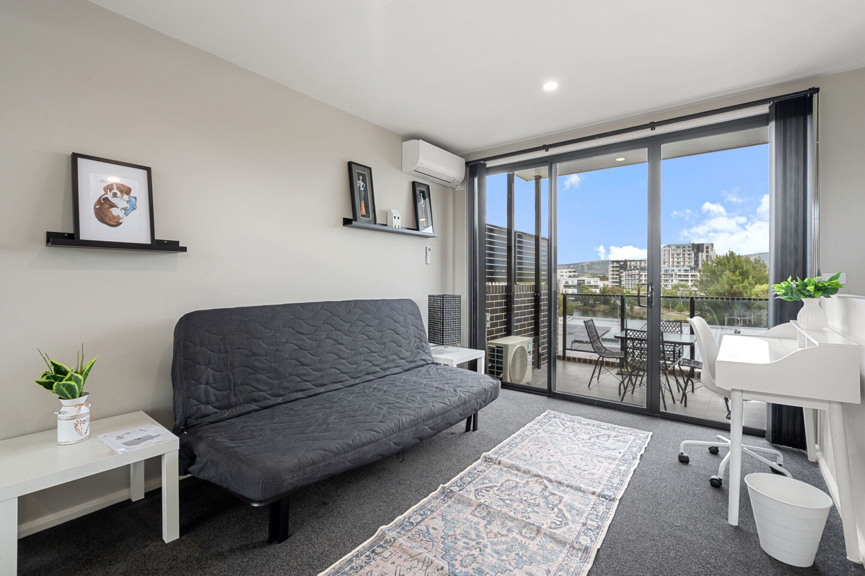 A modern living room with a grey sofa, a white desk, and a large window offering a city view.