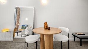 Una habitación moderna con una mesa redonda de madera, dos sillas blancas, una mesita auxiliar y un espejo que refleja una habitación con pared de ladrillo y un cuadro.