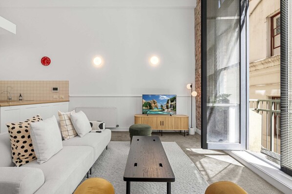 A modern living room with a sofa, ottoman, and a wooden table. There is a TV on a wooden cabinet and a window with blinds. - Classic 1-bed City Stay Near Eateries and Laneways (Melbourne)