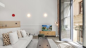 Una sala moderna con un sofá, un reposapiés y una mesa de madera. Hay un televisor sobre un mueble de madera, y una ventana con persianas.