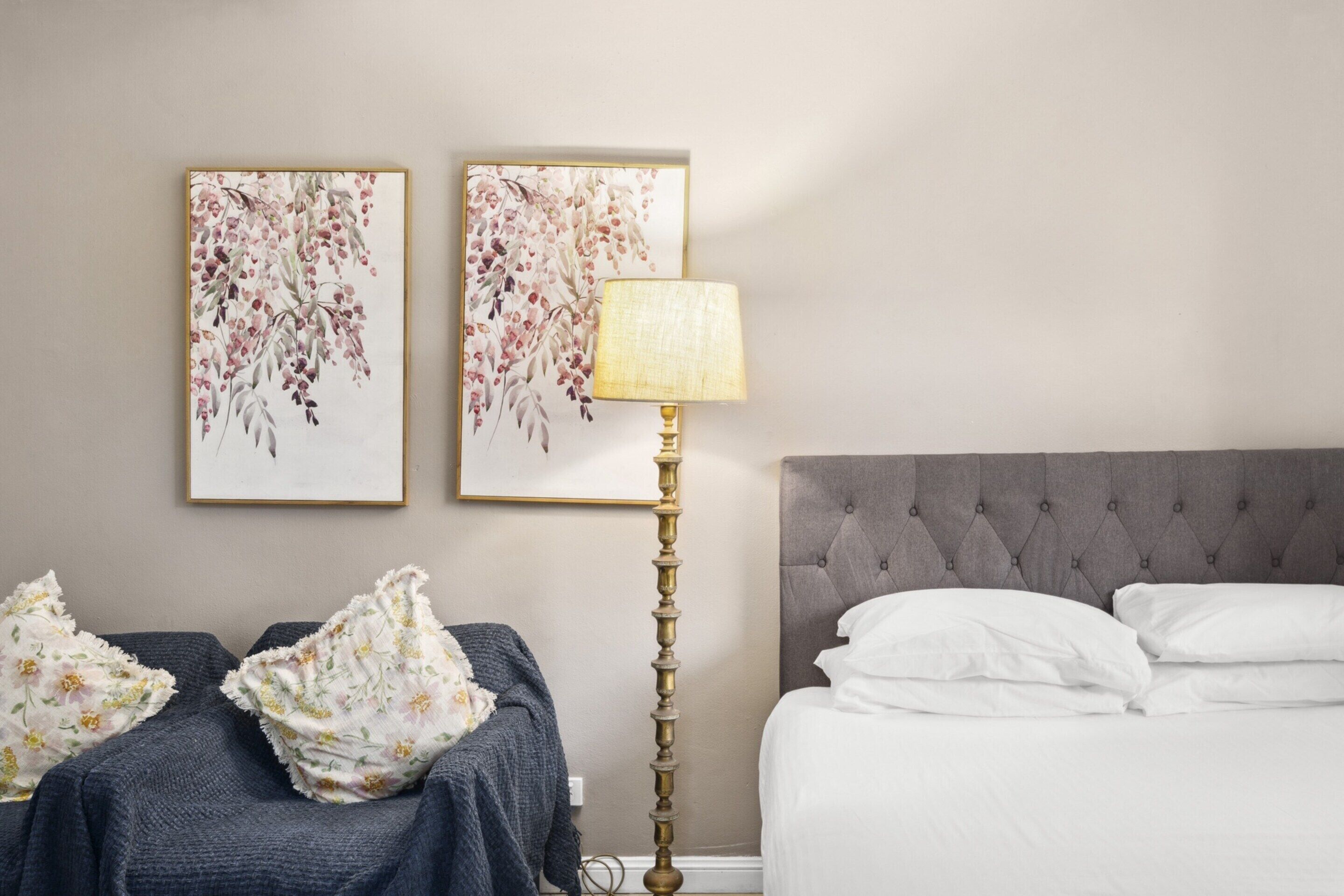 Ein Schlafzimmer mit einem Bett, zwei an der Wand befestigten floralen Bildern, einer Stehlampe und einem grauen, bezogenen Kopfstück.