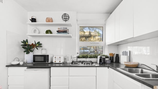 A modern kitchen with white cabinetry, a black countertop, a microwave, a kettle, and a window with a view of a fence and trees. - Chic Urban Paddington Escape (Sydney)
