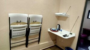 Una habitación pequeña con dos sillas blancas, un estante blanco con una tetera, una mesita con un control remoto y un inodoro morado.
