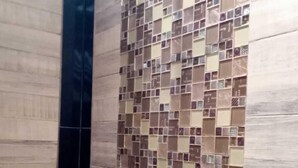 Un baño con pared de ducha azulejada, un panel vertical negro y una repisa metálica con una botella.