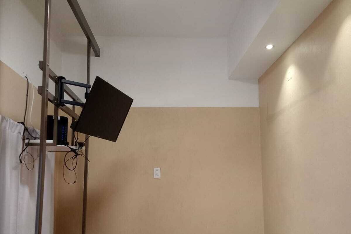 A single bed with a striped blanket, a flat-screen TV mounted on a stand, and a power outlet on the wall.