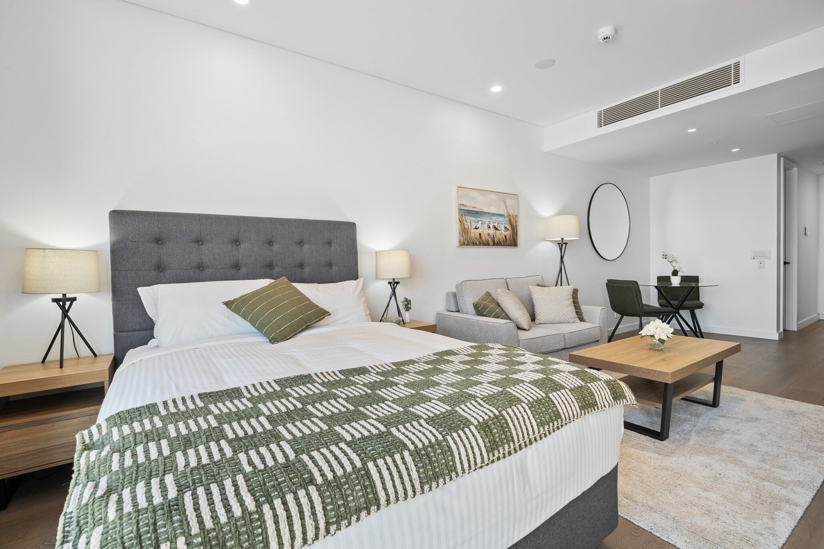 A modern hotel room with a large bed, a sofa, a dining table, and a small desk.