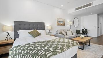 A modern hotel room with a large bed, a sofa, a dining table, and a small desk.