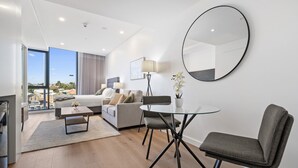 A modern living room with a sofa, a glass coffee table, and a dining table with chairs. There is a large window offering a view of the city. - Bright Studio in Trendy North Fremantle (Perth)