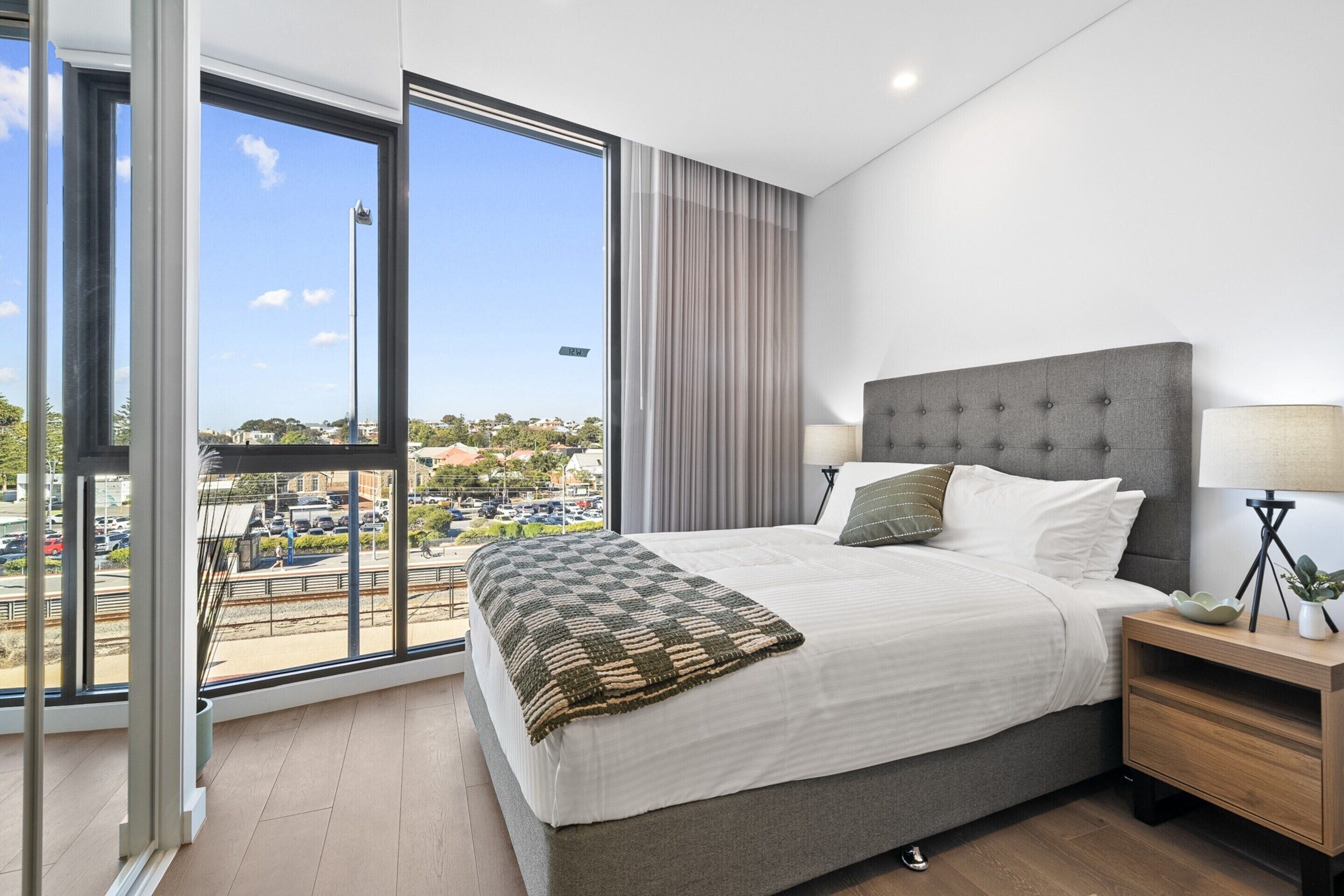 Un dormitorio moderno con una cama grande, una mesita de noche y vistas a un paisaje urbano a través de un ventanal amplio.