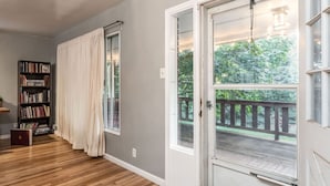 A room with a wooden floor, a bookshelf, a large window with curtains, and a door leading to an outdoor deck. - Cozy, Low-Key, Perfect for Short & Extended Visits (Salt Lake City)