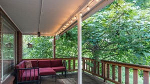 A covered porch with a wooden deck, a red cushioned bench, and a view of lush green trees. - Cozy, Low-Key, Perfect for Short & Extended Visits (Salt Lake City)