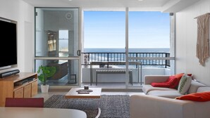 Uma sala de estar moderna com uma TV grande, um sofá, uma mesa de centro e uma varanda com vista para o oceano.