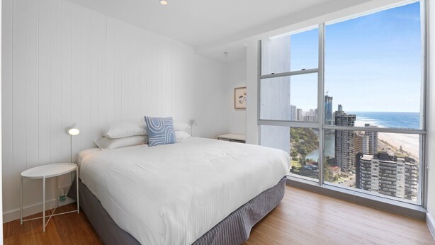 Quarto moderno com cama grande, mesa de cabeceira, luminária e vista para a praia e o horizonte da cidade através de uma janela ampla.