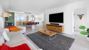 Ein modernes Wohnzimmer mit Sofa, Couchtisch und Flachbildfernseher. Im Hintergrund ist eine Küche mit Essbereich zu sehen.