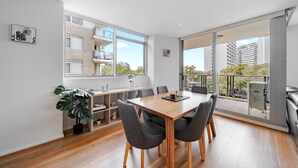 Un espace repas moderne avec une table et des chaises en bois, de grandes fenêtres et une vue sur un immeuble et des arbres.