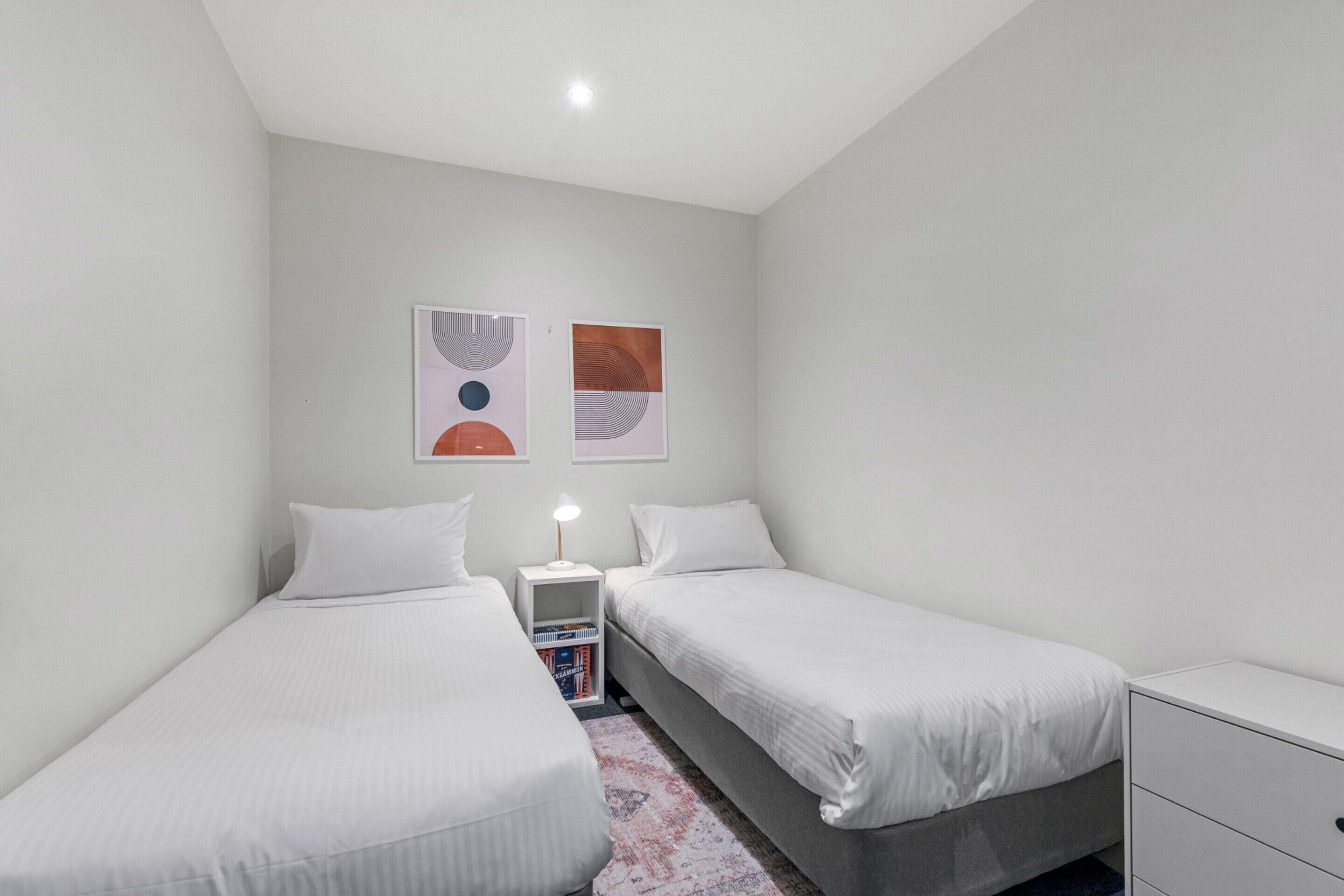 A small, clean room with two single beds, a nightstand with a lamp, and two wall art pieces.