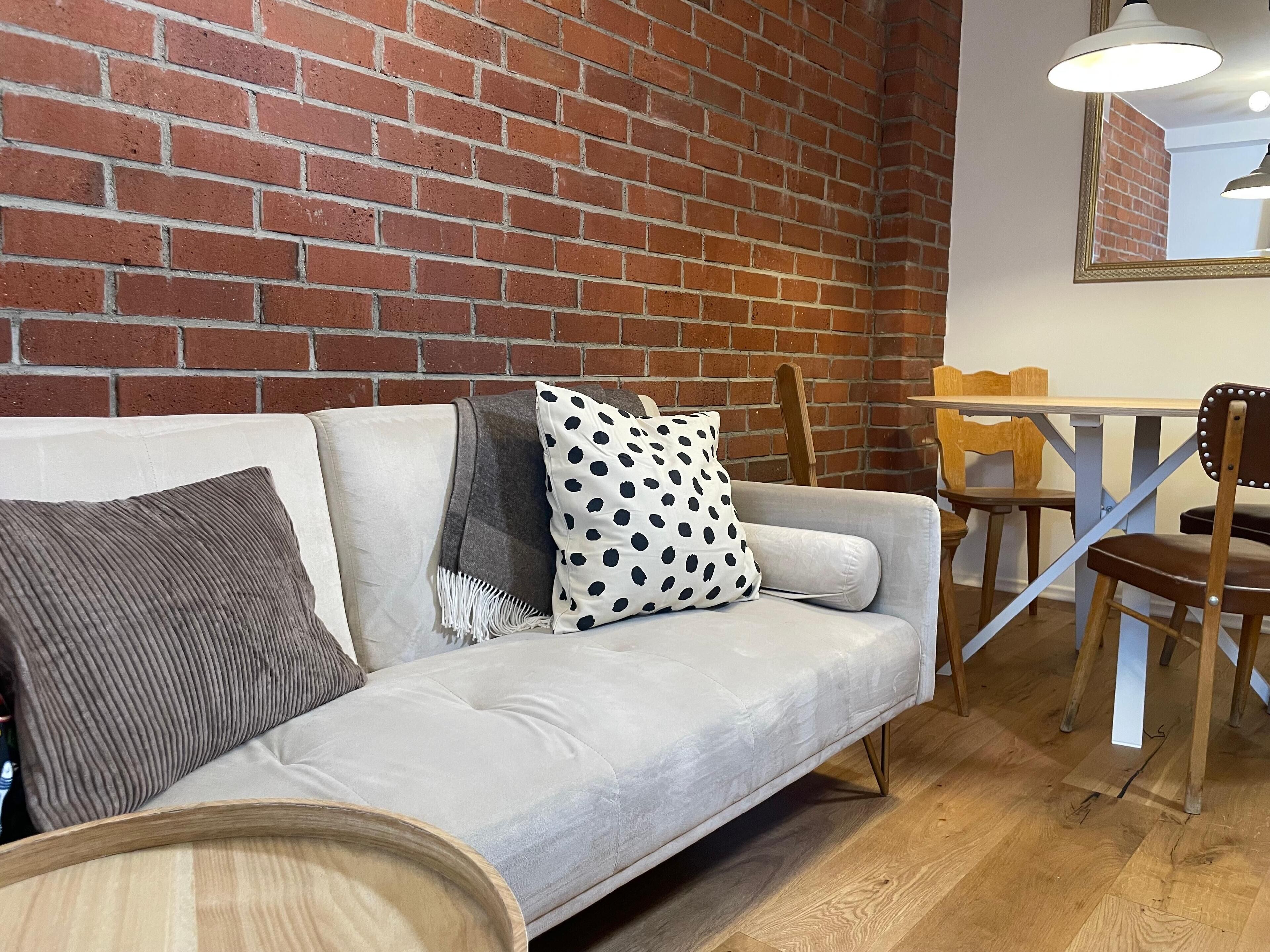 Ein Zimmer mit einer Ziegelwand, einem hellen Sofa mit gemusterten Kissen, einem Holztisch und einem Essbereich mit Stühlen.