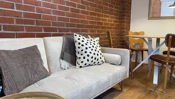 A room with a brick wall, a light-colored sofa with patterned cushions, a wooden coffee table, and a dining area with chairs.