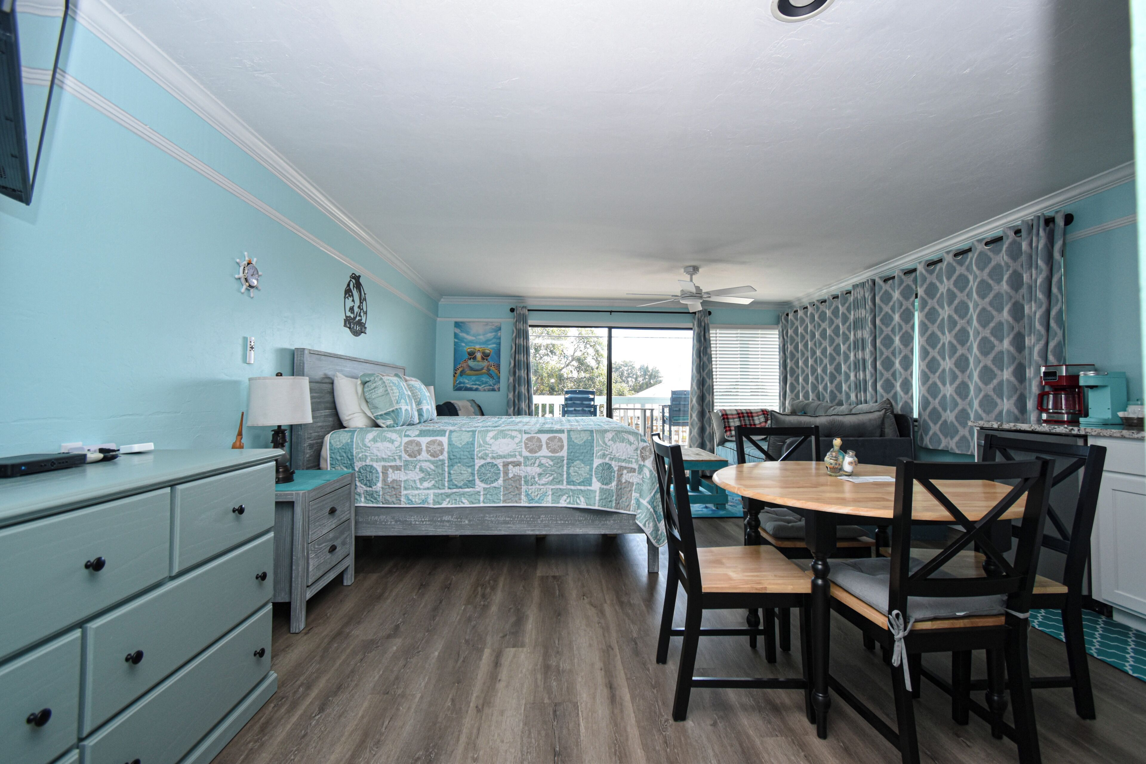 A room with a bed, a dining table with chairs, a dresser, a ceiling fan, and a window with curtains.
