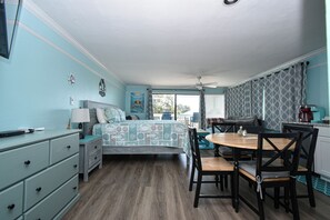 Une pièce comprenant un lit, une table à manger avec des chaises, une commode, un ventilateur de plafond et une fenêtre avec des rideaux.