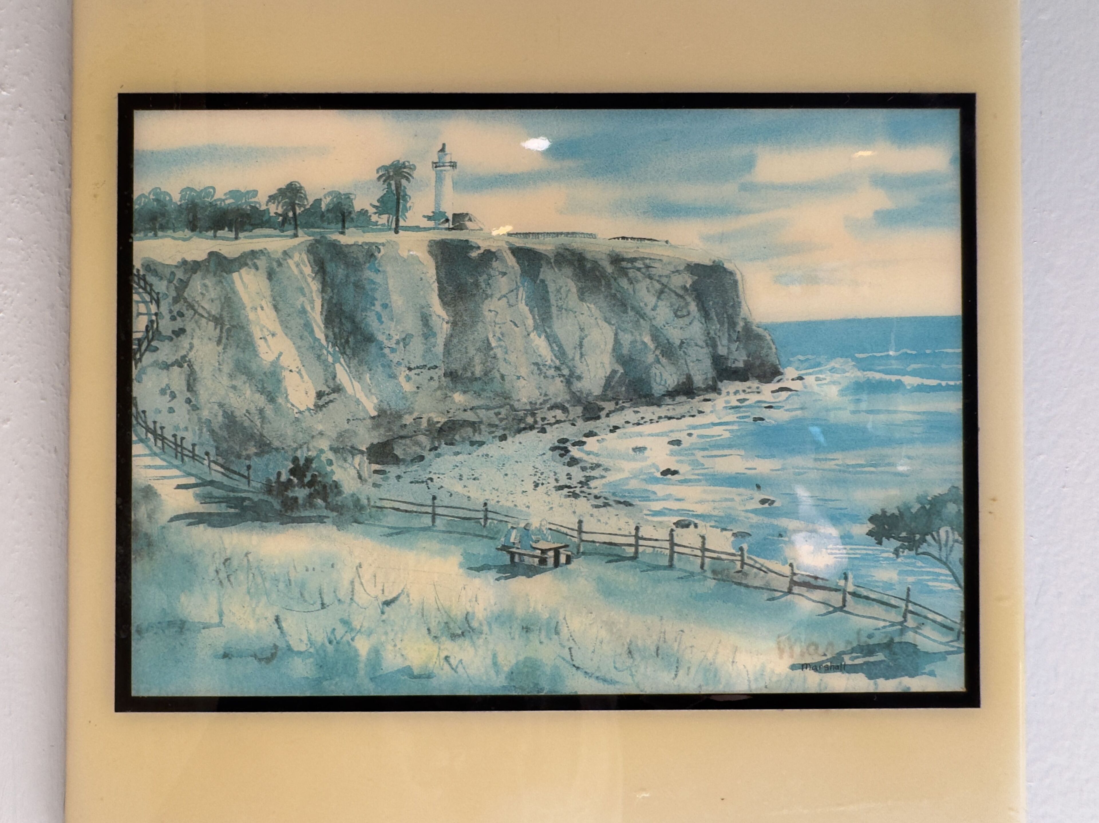 Un quadro ad acquerello incorniciato raffigura una costa con un faro, scogliere e una passerella di legno.