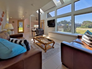 A spacious living room with a sofa, a coffee table, and a TV mounted on the wall. There is a fireplace and a large window offering a view of the outdoors.