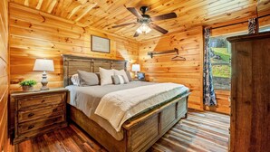 A wooden cabin bedroom with a bed, nightstands, a ceiling fan, and a window with curtains. - Pinot Paradise (Pigeon Forge)