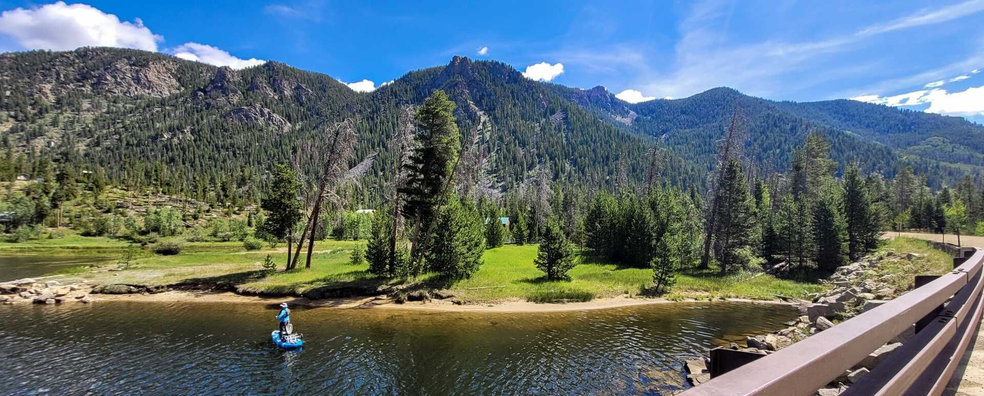 Un río con una persona en una tabla de remo, rodeado de montañas boscosas y un cielo despejado.