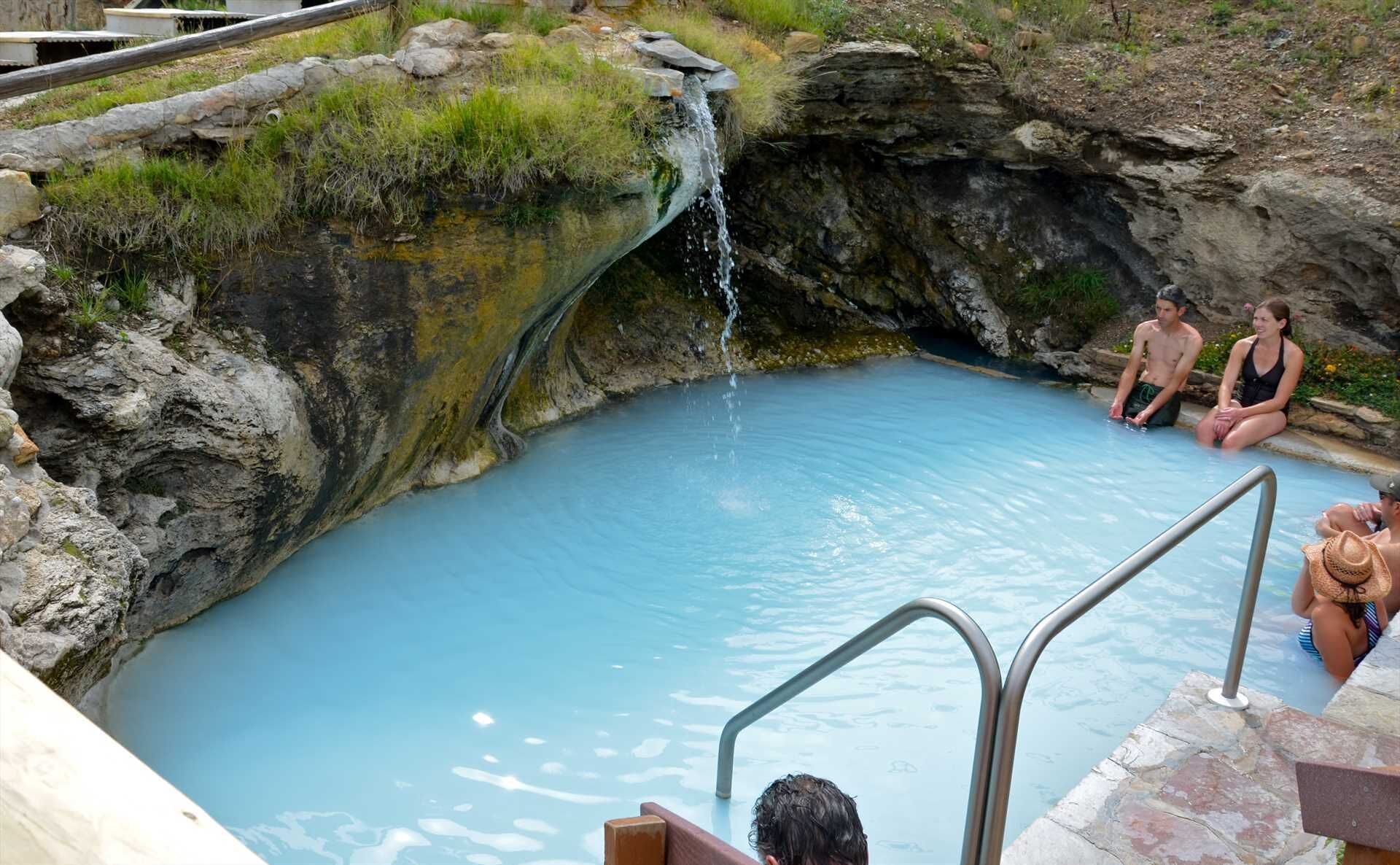 Un manantial termal natural con una pequeña cascada, rodeado de terreno rocoso y vegetación.