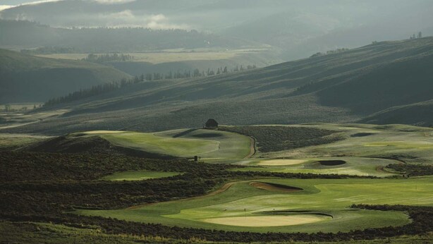Et panorama over en golfbane med bløde bakker, en mindre bygning og fjerne bjerge.