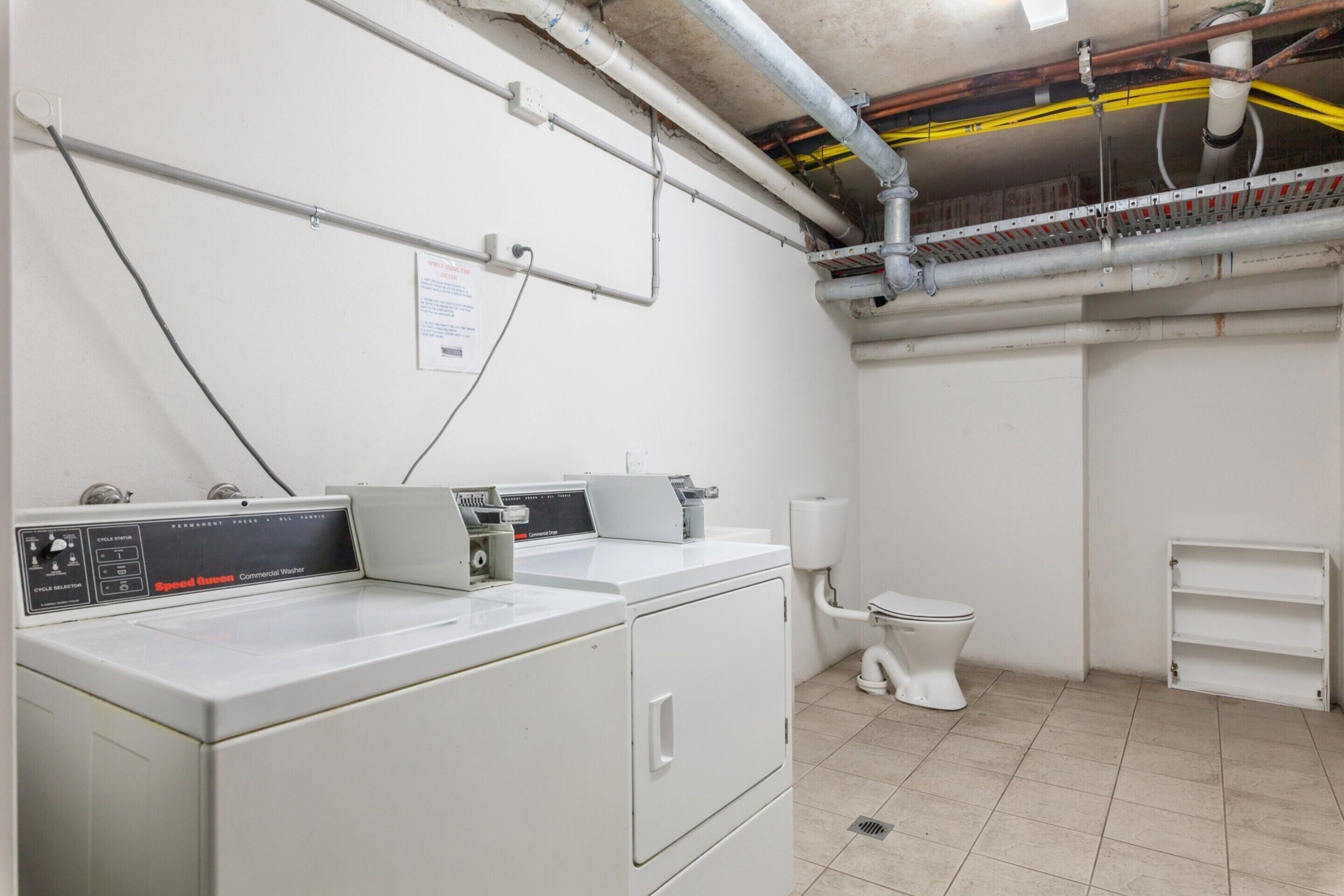 A laundry room with a front-loading commercial washer, a toilet, and a metal shelf unit.