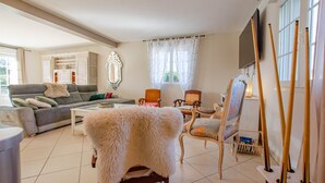 Ein Wohnzimmer mit einem grauen Sofa, einem weißen, flauschigen Teppich und einem Holztisch. Zwei Sessel sind vorhanden, einer mit einem Blumenkissen und der andere mit einem schlichten Kissen. Ein Fernseher ist an der Wand montiert, und mehrere Holzstämme lehnen an der Wand.