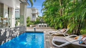 A swimming pool with lounge chairs and a table, surrounded by lush greenery and a modern building. - Riverstone Noosaville 2 Noosaville (Noosaville)