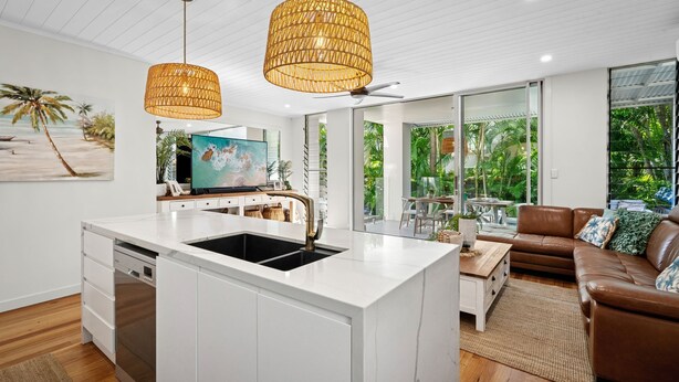 A modern kitchen with a white island, a dining area with a brown leather couch, and large windows offering a view of greenery. - Riverstone Noosaville 2 Noosaville (Noosaville)