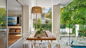 A modern dining area with a wooden table and white chairs, a woven pendant light, and a view of a lush garden and pool. - Riverstone Noosaville 2 Noosaville (Noosaville)