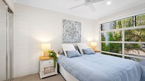 A bedroom with a bed, bedside tables, a ceiling fan, and a window with a view of greenery. - Riverstone Noosaville 2 Noosaville (Noosaville)
