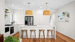 A modern kitchen with white cabinetry, a stainless steel refrigerator, a built-in oven, and a central island with bar stools. - Riverstone Noosaville 2 Noosaville (Noosaville)