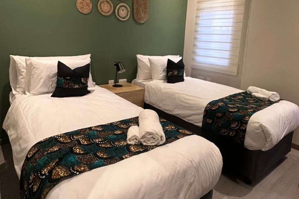 Une chambre d’hôtel avec deux lits simples, chacun agrémenté de draps blancs et de coussins décoratifs. On y trouve une petite table de chevet avec une lampe, ainsi qu’une fenêtre avec des stores.