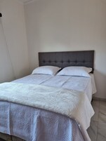Uma cama de casal com cabeceira cinza revestida, roupa de cama branca e parede lisa.