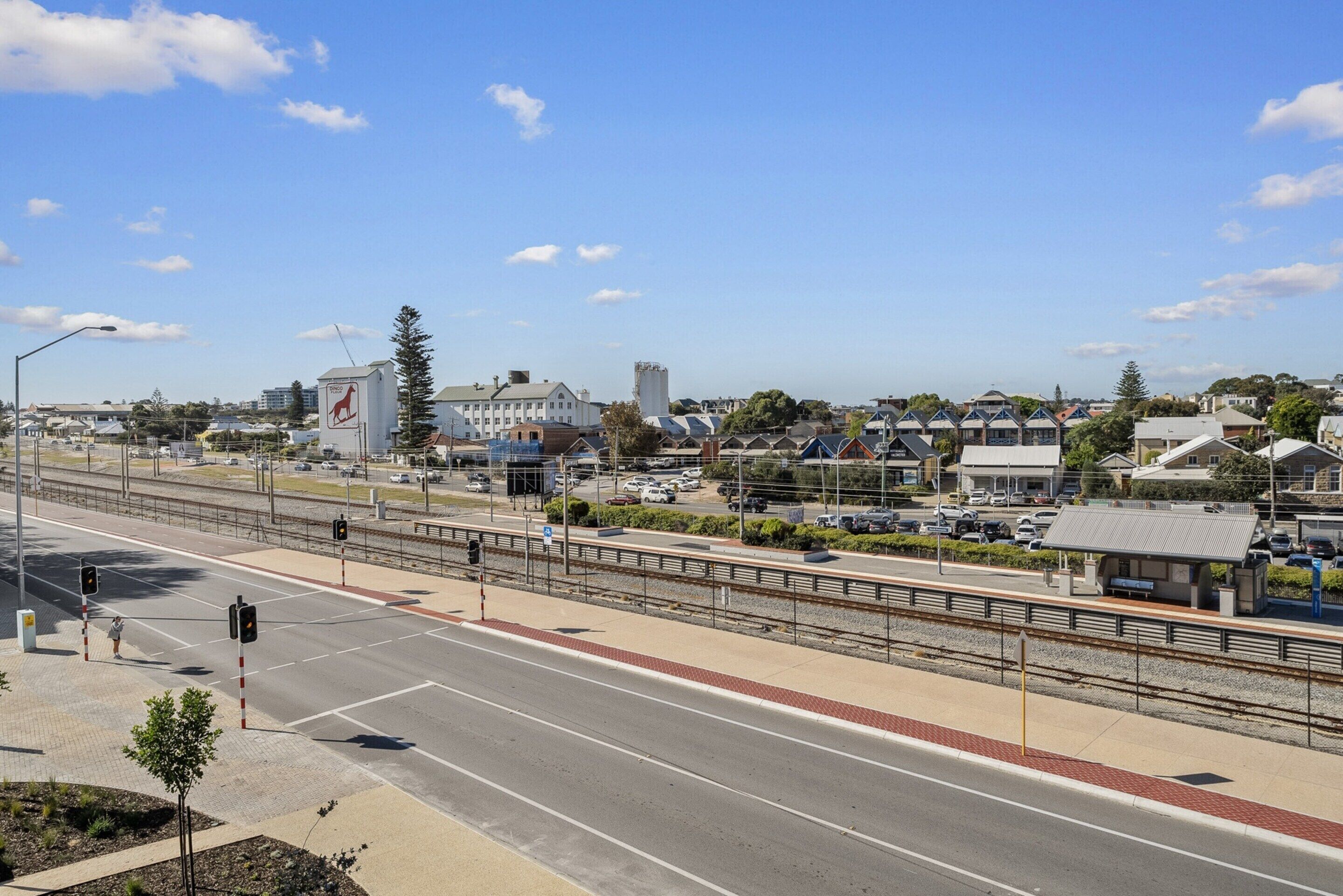 Uma passagem de ferrovia com uma ponte para pedestres, uma rua com calçada e edifícios ao fundo.