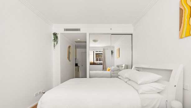 A modern bedroom with a large bed, a bedside table, a mirror, and a small plant.