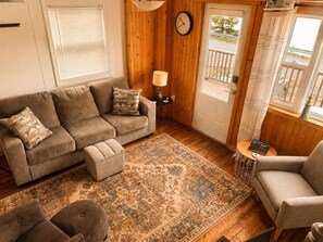 A cozy living room with a sofa, ottoman, and armchair, a patterned rug, a clock, and a door with a balcony view.