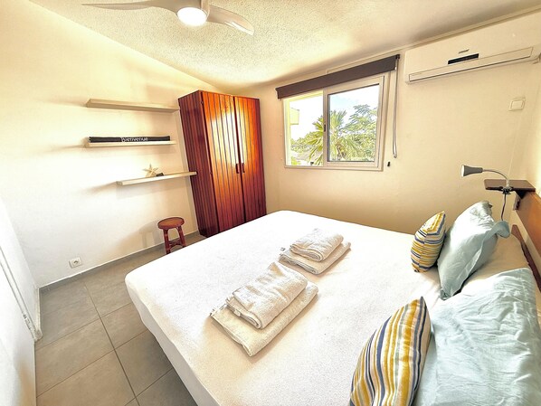 A bedroom with a large bed, a window with a view, a ceiling fan, and a small stool.