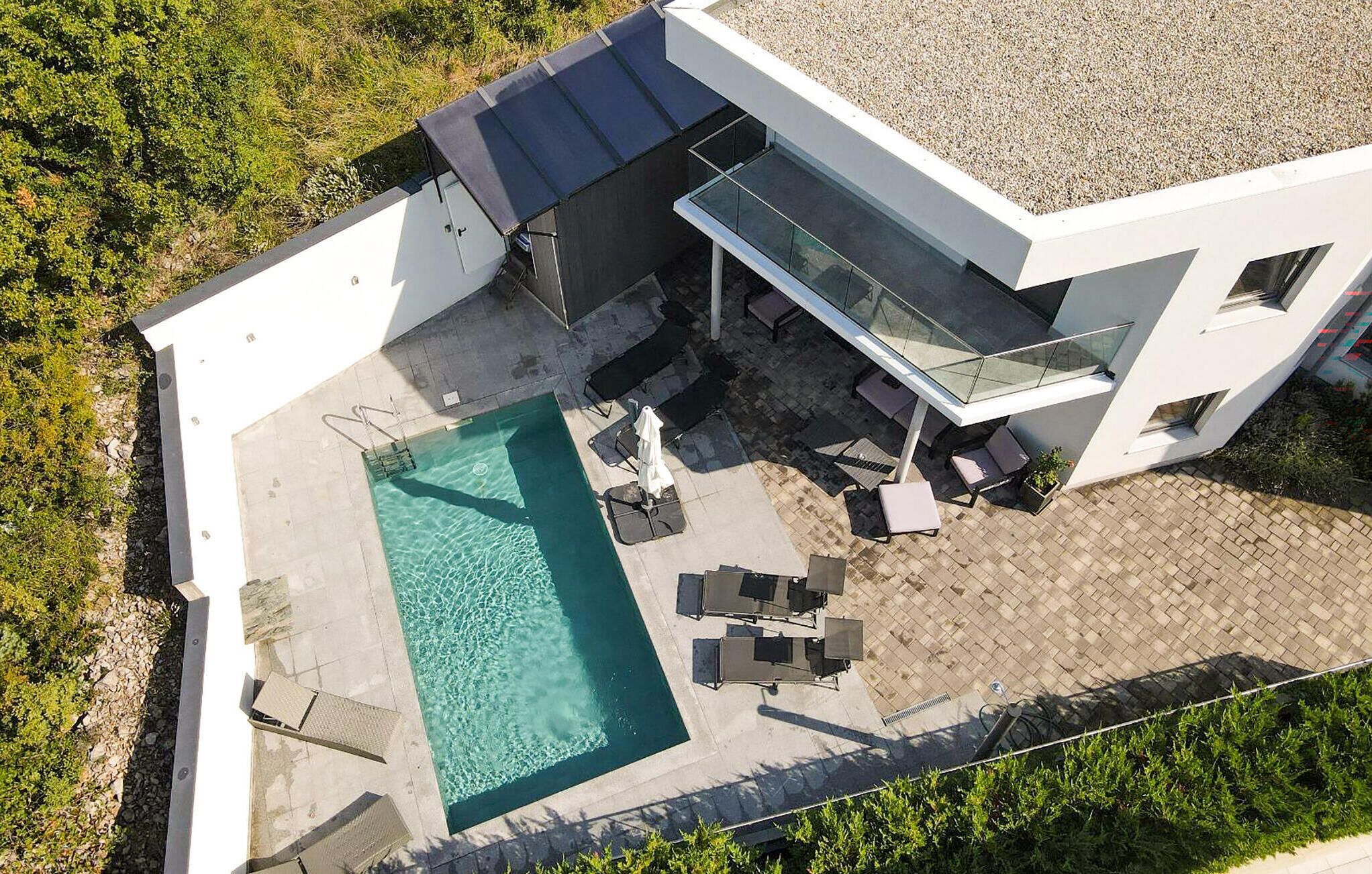 Una casa moderna con piscina, mobiliario exterior y un balcón con paredes de vidrio.