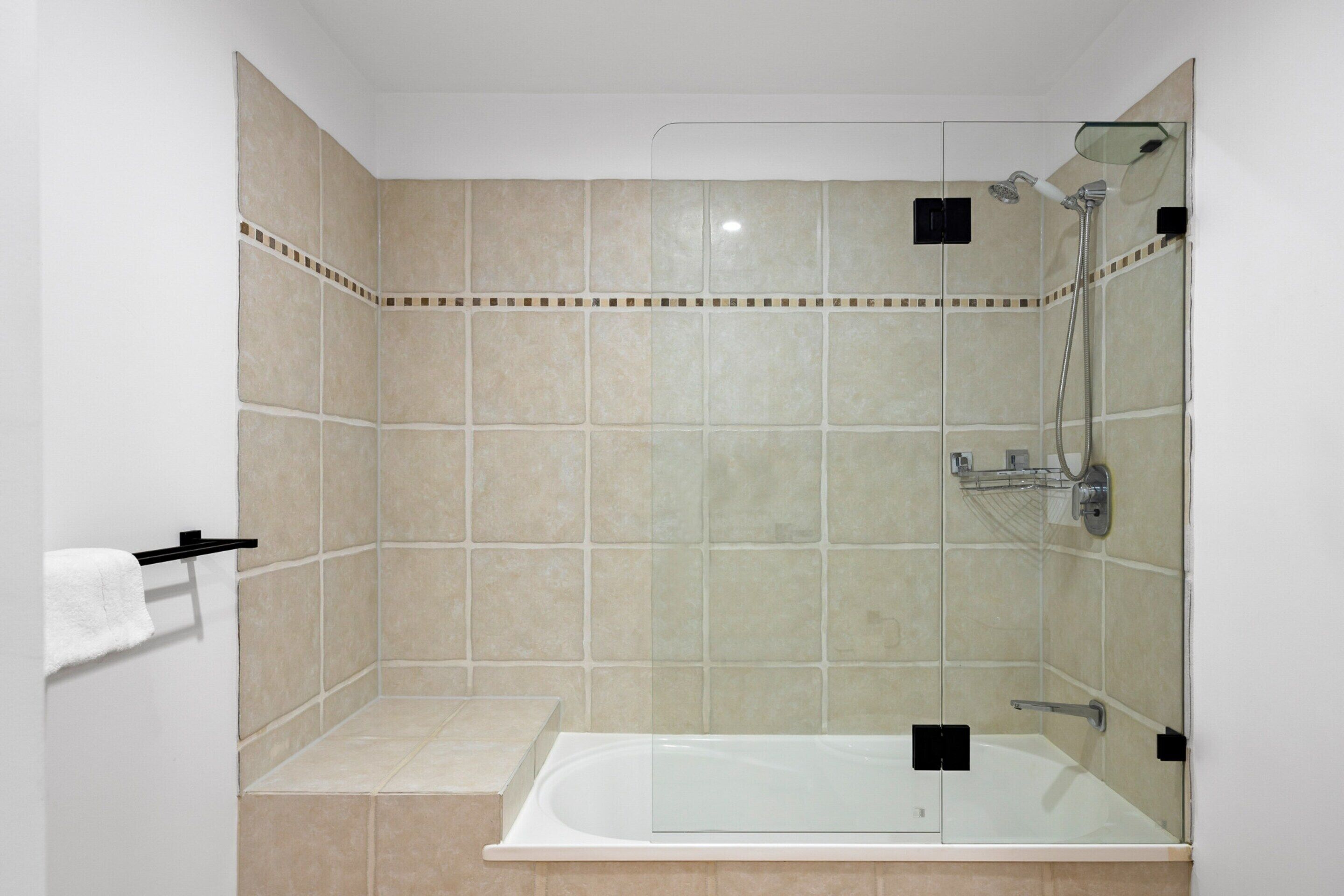 Een badkamer met een douche met glazen wand, een witte badkuip en beige tegels.