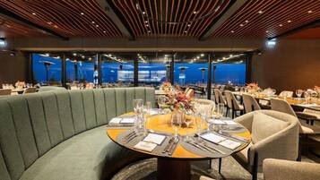 A restaurant with a curved green booth, round tables with chairs, and a view of the cityscape through large windows.