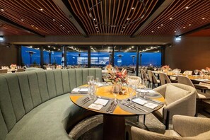 Un restaurante con una mesa semicircular tapizada de verde, mesas redondas con sillas y vistas panorámicas de la ciudad a través de amplias ventanas.