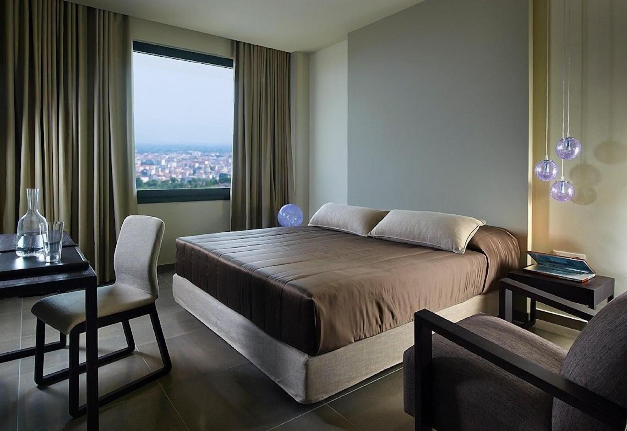 A hotel room with a large bed, a desk, and a chair. There is a view of a cityscape through the window.