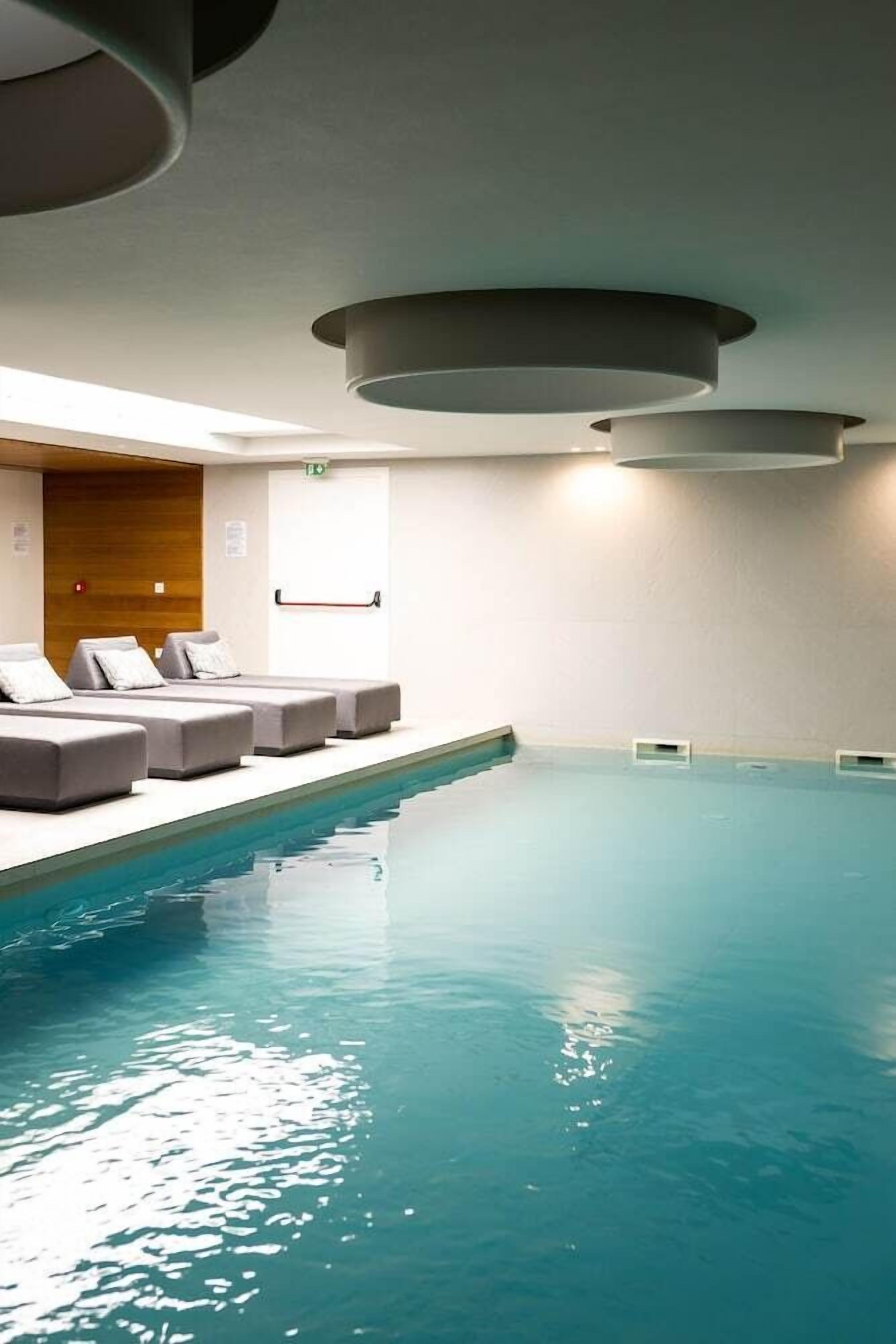 En swimmingpool med et solbelyst interiør, en trævæg og en række grå liggestole.