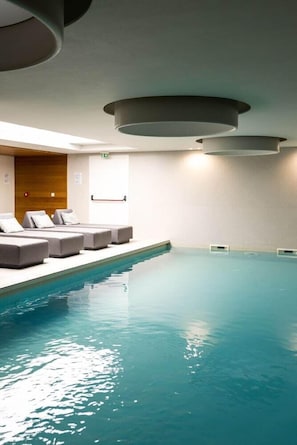 A swimming pool with a sunlit interior, a wooden wall, and a row of grey lounge chairs. - Ananti Resort & Spa (Trikala)